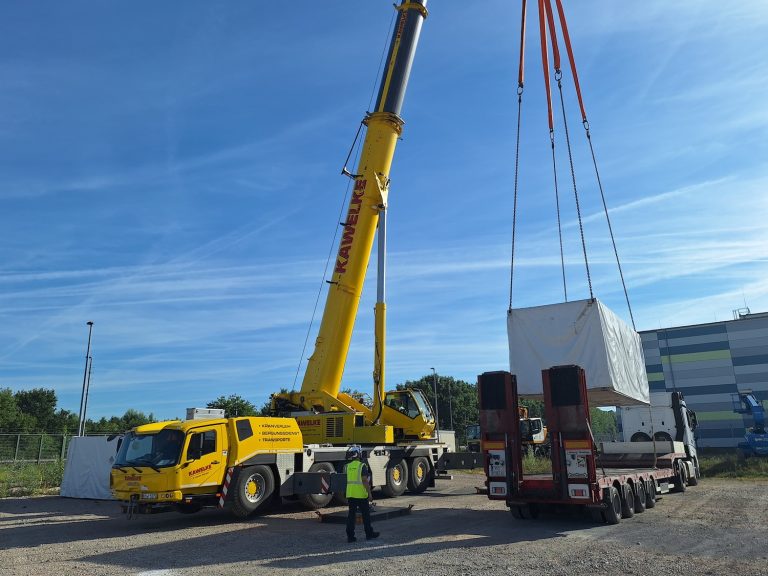 Ein Autokran hebt einen großen, weiß verpackten Batteriespeicher von einem Tieflader auf einer Baustelle im Energiepark Philippsburg. Im Vordergrund überwacht eine Person in Warnweste den Hebevorgang. Im Hintergrund sind ein Industriegebäude und weitere Baustellenfahrzeuge unter blauem Himmel zu sehen.