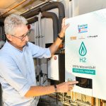 Ein Mann steht in einem Heizungskeller vor einer wandhängenden Wasserstoff-Heizung und bedient das Display des Geräts. Auf der Front ist der Hinweis „H2 – 100 % Wasserstoff“ zu sehen. Die Anlage ist Teil eines Projekts zur Versorgung von Gebäuden mit Wasserstoff über das bestehende Gasnetz. Im Hintergrund sind Rohrleitungen und weitere Komponenten der Heizungsinstallation erkennbar.