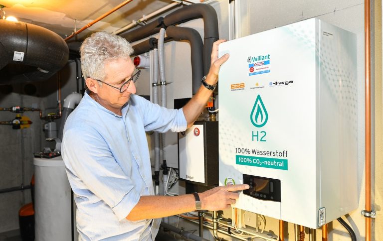 Ein Mann steht in einem Heizungskeller vor einer wandhängenden Wasserstoff-Heizung und bedient das Display des Geräts. Auf der Front ist der Hinweis „H2 – 100 % Wasserstoff“ zu sehen. Die Anlage ist Teil eines Projekts zur Versorgung von Gebäuden mit Wasserstoff über das bestehende Gasnetz. Im Hintergrund sind Rohrleitungen und weitere Komponenten der Heizungsinstallation erkennbar.