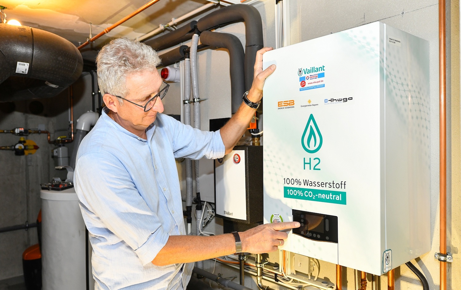 Ein Mann steht in einem Heizungskeller vor einer wandhängenden Wasserstoff-Heizung und bedient das Display des Geräts. Auf der Front ist der Hinweis „H2 – 100 % Wasserstoff“ zu sehen. Die Anlage ist Teil eines Projekts zur Versorgung von Gebäuden mit Wasserstoff über das bestehende Gasnetz. Im Hintergrund sind Rohrleitungen und weitere Komponenten der Heizungsinstallation erkennbar.