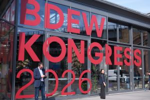 Außenansicht des BDEW-Kongresses 2026: Die Glasfassade des Veranstaltungsgebäudes ist mit großformatigen roten Buchstaben beschriftet, die den Schriftzug „BDEW Kongress 2026“ bilden. Vor dem Eingang stehen mehrere Teilnehmende, während sich die umliegende Architektur und der Himmel in den Glasflächen spiegeln.
