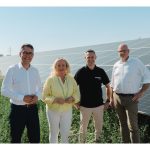 Vier Personen stehen vor einer großflächigen Photovoltaikanlage und blicken in die Kamera. Im Hintergrund erstrecken sich Reihen von Solarmodulen unter einem klaren blauen Himmel. Die Gruppe steht auf einer begrünten Freifläche vor den Modulen und symbolisiert den Ausbau erneuerbarer Energien.