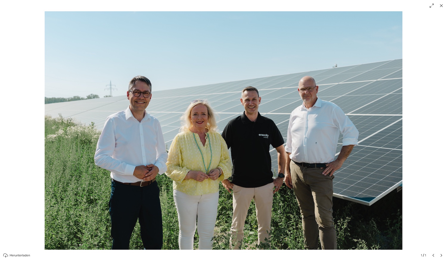 Vier Personen stehen vor einer großflächigen Photovoltaikanlage und blicken in die Kamera. Im Hintergrund erstrecken sich Reihen von Solarmodulen unter einem klaren blauen Himmel. Die Gruppe steht auf einer begrünten Freifläche vor den Modulen und symbolisiert den Ausbau erneuerbarer Energien.