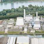 Luftaufnahme eines Energie- und Fernwärmestandorts am Kölner Hafen. Die Anlage liegt auf einer Landzunge zwischen einem Hafenbecken und dem Rhein. Zu sehen sind mehrere Industriegebäude, ein großer Kühlturm, Schornsteine, Rohrleitungen sowie runde Speicher- und Tankanlagen. Im Hintergrund fließt der Rhein, dahinter erstrecken sich Grünflächen und Bäume. Das Bild zeigt die bestehende Infrastruktur des Standorts Köln-Niehl, an dem eine große Flusswasser-Wärmepumpe errichtet werden soll.