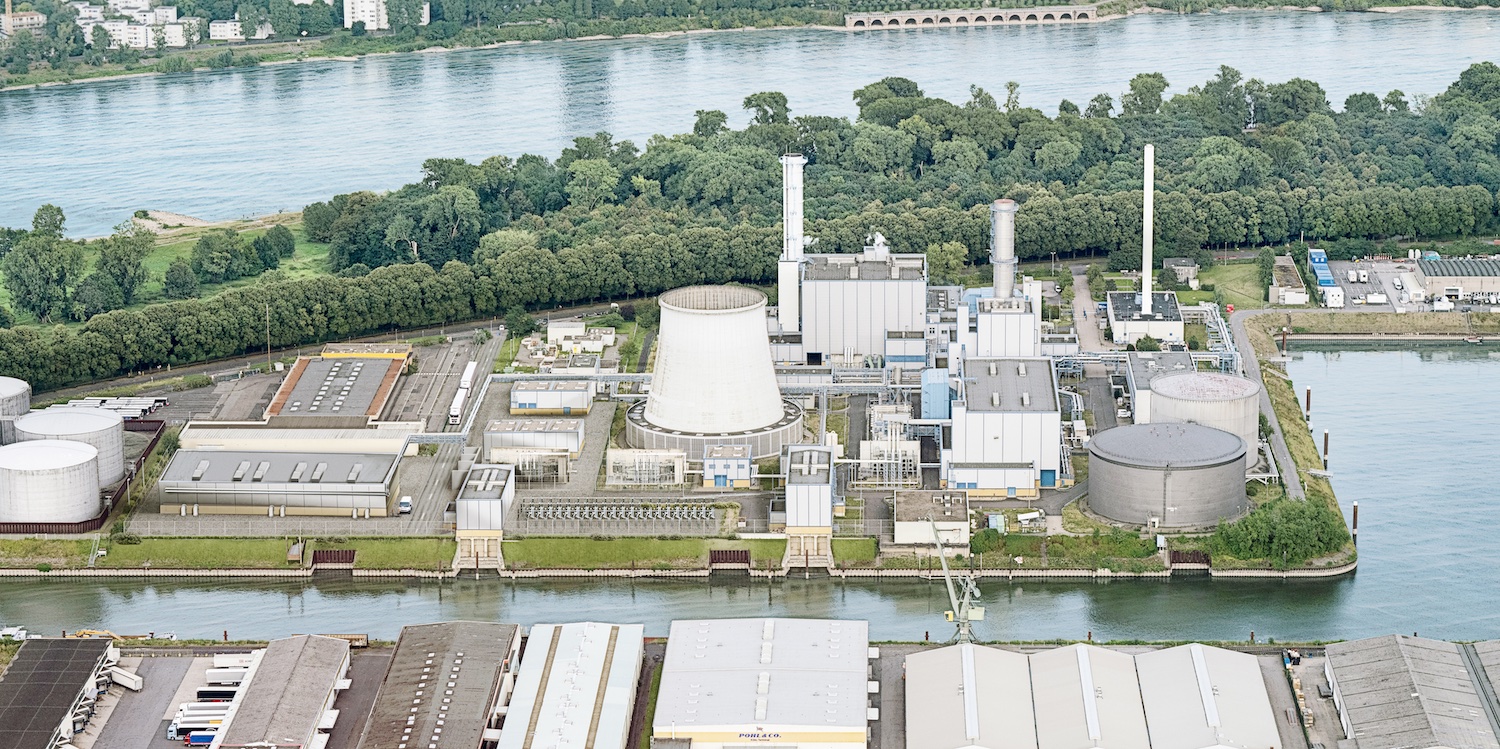 Luftaufnahme eines Energie- und Fernwärmestandorts am Kölner Hafen. Die Anlage liegt auf einer Landzunge zwischen einem Hafenbecken und dem Rhein. Zu sehen sind mehrere Industriegebäude, ein großer Kühlturm, Schornsteine, Rohrleitungen sowie runde Speicher- und Tankanlagen. Im Hintergrund fließt der Rhein, dahinter erstrecken sich Grünflächen und Bäume. Das Bild zeigt die bestehende Infrastruktur des Standorts Köln-Niehl, an dem eine große Flusswasser-Wärmepumpe errichtet werden soll.