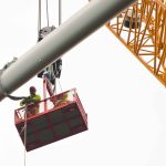 Zwei Monteure in Schutzkleidung stehen auf einer angehobenen Arbeitsplattform und begleiten die Montage eines Rotorblatts einer Windkraftanlage. Das lange Rotorblatt wird von einem großen Kran in Position gebracht. Rechts im Bild ist der gelbe Ausleger des Krans zu sehen. Der helle, bedeckte Himmel bildet den Hintergrund der Arbeiten in großer Höhe.