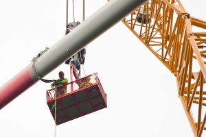 Zwei Monteure in Schutzkleidung stehen auf einer angehobenen Arbeitsplattform und begleiten die Montage eines Rotorblatts einer Windkraftanlage. Das lange Rotorblatt wird von einem großen Kran in Position gebracht. Rechts im Bild ist der gelbe Ausleger des Krans zu sehen. Der helle, bedeckte Himmel bildet den Hintergrund der Arbeiten in großer Höhe.