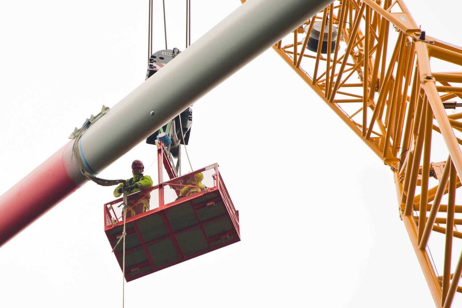 Zwei Monteure in Schutzkleidung stehen auf einer angehobenen Arbeitsplattform und begleiten die Montage eines Rotorblatts einer Windkraftanlage. Das lange Rotorblatt wird von einem großen Kran in Position gebracht. Rechts im Bild ist der gelbe Ausleger des Krans zu sehen. Der helle, bedeckte Himmel bildet den Hintergrund der Arbeiten in großer Höhe.