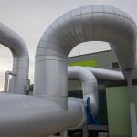 Gedämmte Fernwärmeleitungen verlaufen in mehreren Bögen über das Außengelände einer Energieanlage. Die groß dimensionierten silberfarbenen Rohre führen entlang eines modernen Technikgebäudes und veranschaulichen die Infrastruktur eines Wärmenetzes.