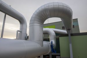 Gedämmte Fernwärmeleitungen verlaufen in mehreren Bögen über das Außengelände einer Energieanlage. Die groß dimensionierten silberfarbenen Rohre führen entlang eines modernen Technikgebäudes und veranschaulichen die Infrastruktur eines Wärmenetzes.