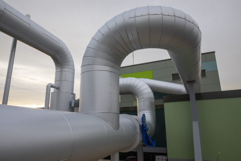 Gedämmte Fernwärmeleitungen verlaufen in mehreren Bögen über das Außengelände einer Energieanlage. Die groß dimensionierten silberfarbenen Rohre führen entlang eines modernen Technikgebäudes und veranschaulichen die Infrastruktur eines Wärmenetzes.