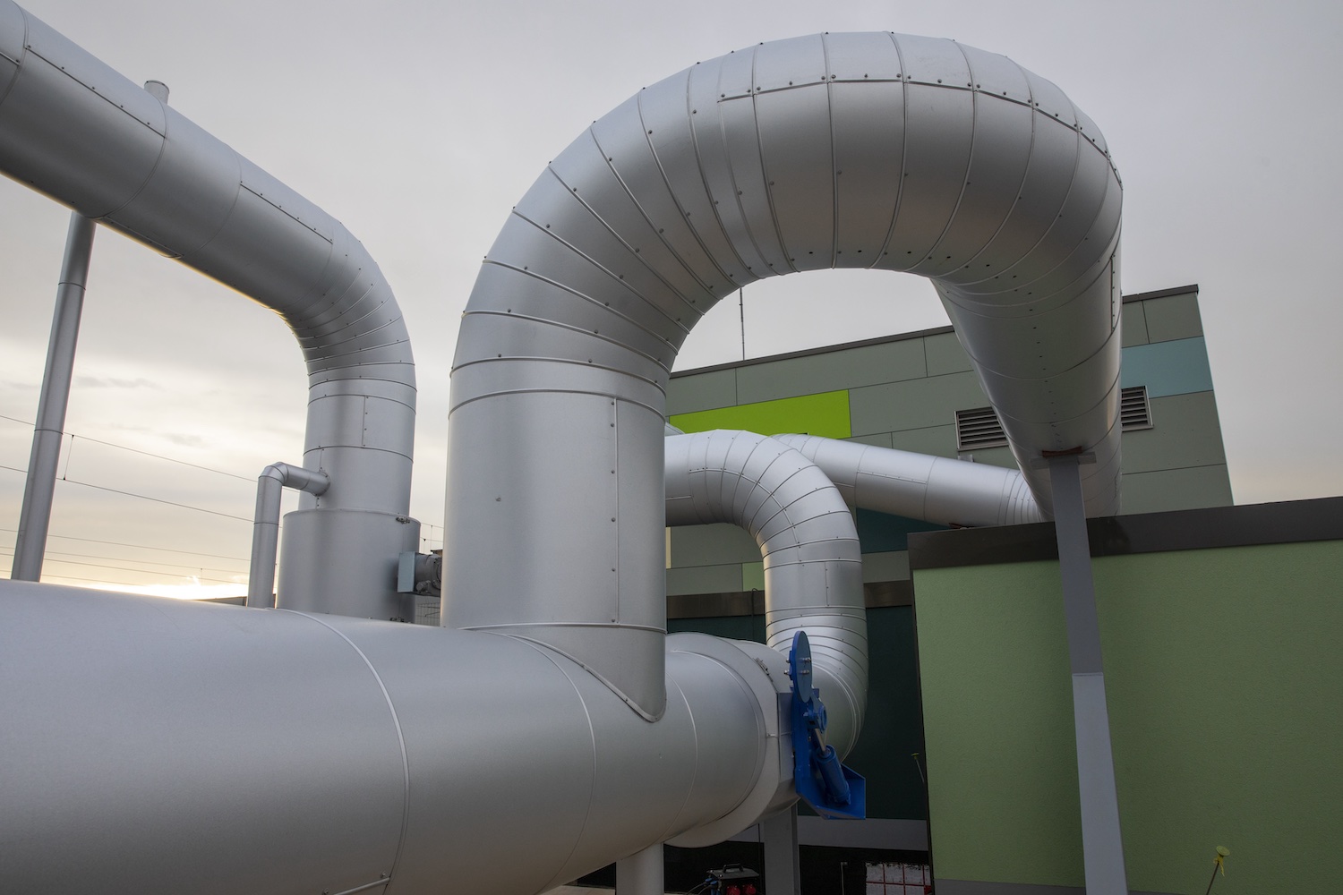 Gedämmte Fernwärmeleitungen verlaufen in mehreren Bögen über das Außengelände einer Energieanlage. Die groß dimensionierten silberfarbenen Rohre führen entlang eines modernen Technikgebäudes und veranschaulichen die Infrastruktur eines Wärmenetzes.
