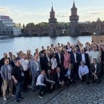 Gruppenfoto des GWAdriga-Teams am Ufer der Spree in Berlin. Rund 70 Mitarbeitende und Partner stehen und knien auf einer gepflasterten Uferpromenade und blicken in die Kamera. Im Hintergrund ist die historische Oberbaumbrücke mit ihren markanten Türmen zu sehen.