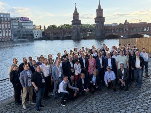 Gruppenfoto des GWAdriga-Teams am Ufer der Spree in Berlin. Rund 70 Mitarbeitende und Partner stehen und knien auf einer gepflasterten Uferpromenade und blicken in die Kamera. Im Hintergrund ist die historische Oberbaumbrücke mit ihren markanten Türmen zu sehen.