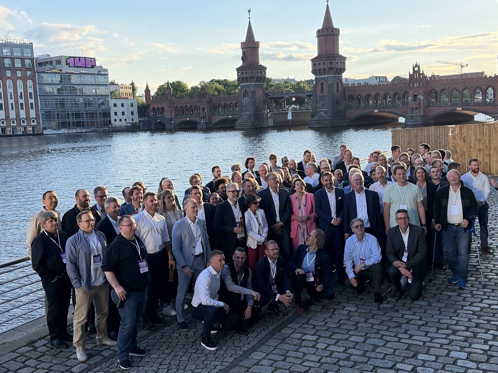 Gruppenfoto des GWAdriga-Teams am Ufer der Spree in Berlin. Rund 70 Mitarbeitende und Partner stehen und knien auf einer gepflasterten Uferpromenade und blicken in die Kamera. Im Hintergrund ist die historische Oberbaumbrücke mit ihren markanten Türmen zu sehen.