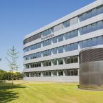 Außenansicht der RheinEnergie-Unternehmenszentrale: Das moderne, mehrgeschossige Bürogebäude mit heller Metall-Glas-Fassade trägt gut sichtbar den Schriftzug „RheinEnergie“. Im Vordergrund liegen eine gepflegte Rasenfläche und ein zylindrischer Lüftungsbau, links verläuft ein Fuß- und Radweg entlang einer Baumreihe. Der Himmel ist wolkenlos und blau.