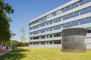 Außenansicht der RheinEnergie-Unternehmenszentrale: Das moderne, mehrgeschossige Bürogebäude mit heller Metall-Glas-Fassade trägt gut sichtbar den Schriftzug „RheinEnergie“. Im Vordergrund liegen eine gepflegte Rasenfläche und ein zylindrischer Lüftungsbau, links verläuft ein Fuß- und Radweg entlang einer Baumreihe. Der Himmel ist wolkenlos und blau.