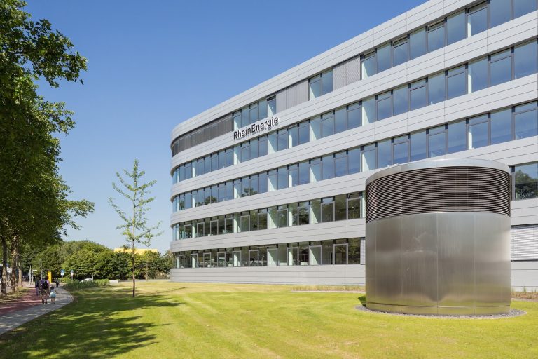 Außenansicht der RheinEnergie-Unternehmenszentrale: Das moderne, mehrgeschossige Bürogebäude mit heller Metall-Glas-Fassade trägt gut sichtbar den Schriftzug „RheinEnergie“. Im Vordergrund liegen eine gepflegte Rasenfläche und ein zylindrischer Lüftungsbau, links verläuft ein Fuß- und Radweg entlang einer Baumreihe. Der Himmel ist wolkenlos und blau.