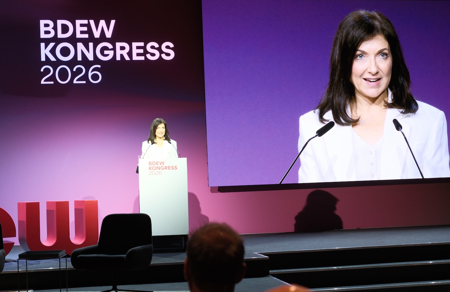 Bundeswirtschaftsministerin Katharina Reiche spricht auf dem BDEW-Kongress 2026 an einem Rednerpult vor einem großen Publikum. Hinter ihr zeigt eine überdimensionale Leinwand eine Nahaufnahme der Ministerin während ihrer Rede. Der Bühnenhintergrund ist in violetten und magentafarbenen Tönen gestaltet und trägt das Logo „BDEW Kongress 2026“.