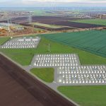 Luftaufnahme einer ländlich geprägten Landschaft mit Feldern, Windkraftanlagen und einem Umspannwerk. In die Aufnahme ist eine Visualisierung eines großflächigen Batteriespeichers eingebettet: Mehrere Reihen von Speichercontainern und technische Anlagen sind auf zwei Arealen dargestellt, die über Straßen miteinander verbunden sind. Das Bild zeigt die geplante Integration eines Batterie-Großspeichers in die bestehende Energieinfrastruktur zur Speicherung und Bereitstellung von erneuerbar erzeugtem Strom.