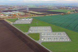 Luftaufnahme einer ländlich geprägten Landschaft mit Feldern, Windkraftanlagen und einem Umspannwerk. In die Aufnahme ist eine Visualisierung eines großflächigen Batteriespeichers eingebettet: Mehrere Reihen von Speichercontainern und technische Anlagen sind auf zwei Arealen dargestellt, die über Straßen miteinander verbunden sind. Das Bild zeigt die geplante Integration eines Batterie-Großspeichers in die bestehende Energieinfrastruktur zur Speicherung und Bereitstellung von erneuerbar erzeugtem Strom.