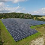 Großflächiger Solarpark mit zahlreichen Photovoltaikmodulen auf einer begrünten Freifläche am Waldrand. Im Hintergrund stehen drei Windkraftanlagen auf einem bewaldeten Höhenzug unter einem teilweise bewölkten Himmel. Die Aufnahme zeigt die Kombination von Solar- und Windenergie in einer ländlichen Landschaft.