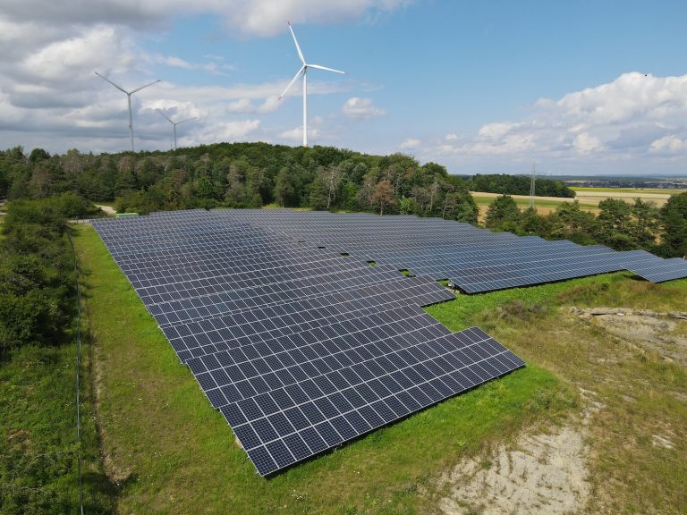 Großflächiger Solarpark mit zahlreichen Photovoltaikmodulen auf einer begrünten Freifläche am Waldrand. Im Hintergrund stehen drei Windkraftanlagen auf einem bewaldeten Höhenzug unter einem teilweise bewölkten Himmel. Die Aufnahme zeigt die Kombination von Solar- und Windenergie in einer ländlichen Landschaft.