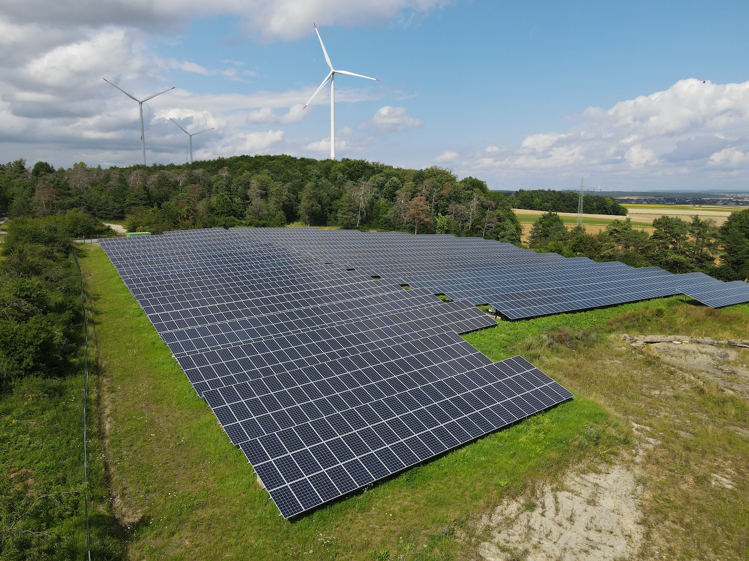 Großflächiger Solarpark mit zahlreichen Photovoltaikmodulen auf einer begrünten Freifläche am Waldrand. Im Hintergrund stehen drei Windkraftanlagen auf einem bewaldeten Höhenzug unter einem teilweise bewölkten Himmel. Die Aufnahme zeigt die Kombination von Solar- und Windenergie in einer ländlichen Landschaft.