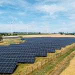 Großflächiger Solarpark auf einer leicht hügeligen Agrarlandschaft. Im Vordergrund erstrecken sich zahlreiche dunkelblaue Photovoltaikmodule, im Hintergrund stehen mehrere Windkraftanlagen auf Feldern unter einem weitläufigen, leicht bewölkten Himmel.