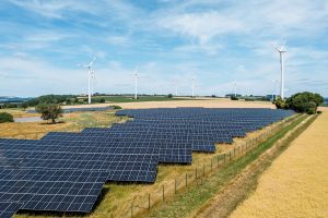 Großflächiger Solarpark auf einer leicht hügeligen Agrarlandschaft. Im Vordergrund erstrecken sich zahlreiche dunkelblaue Photovoltaikmodule, im Hintergrund stehen mehrere Windkraftanlagen auf Feldern unter einem weitläufigen, leicht bewölkten Himmel.