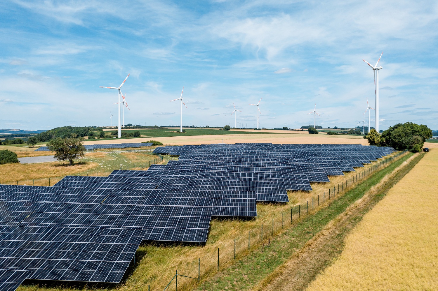 Großflächiger Solarpark auf einer leicht hügeligen Agrarlandschaft. Im Vordergrund erstrecken sich zahlreiche dunkelblaue Photovoltaikmodule, im Hintergrund stehen mehrere Windkraftanlagen auf Feldern unter einem weitläufigen, leicht bewölkten Himmel.