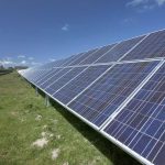 Reihen von Photovoltaikmodulen auf einer Freiflächen-Solaranlage unter blauem Himmel mit einzelnen Wolken. Die Solarpaneele erstrecken sich entlang eines begrünten Geländes und dominieren den Vordergrund. Im Hintergrund sind Windkraftanlagen sowie einige Gebäude zu erkennen.