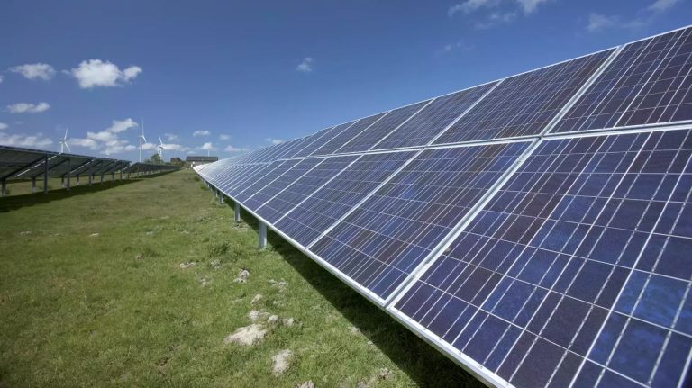 Reihen von Photovoltaikmodulen auf einer Freiflächen-Solaranlage unter blauem Himmel mit einzelnen Wolken. Die Solarpaneele erstrecken sich entlang eines begrünten Geländes und dominieren den Vordergrund. Im Hintergrund sind Windkraftanlagen sowie einige Gebäude zu erkennen.