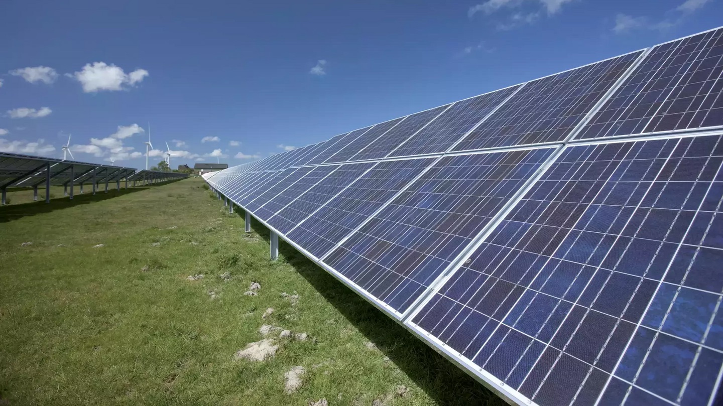 Reihen von Photovoltaikmodulen auf einer Freiflächen-Solaranlage unter blauem Himmel mit einzelnen Wolken. Die Solarpaneele erstrecken sich entlang eines begrünten Geländes und dominieren den Vordergrund. Im Hintergrund sind Windkraftanlagen sowie einige Gebäude zu erkennen.