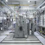 Blick in eine helle Versuchshalle mit einem technisch komplexen Einzylinder-Teststand. Im Zentrum steht eine großformatige Forschungsanlage aus Metall mit zahlreichen Rohrleitungen, Ventilen, Messinstrumenten und Steuerungseinheiten. Die Anlage ist von weiterer Labor- und Prüftechnik umgeben und dient der Untersuchung motorischer Verbrennungsprozesse von Wasserstoff und Ammoniak am WTZ Roßlau.