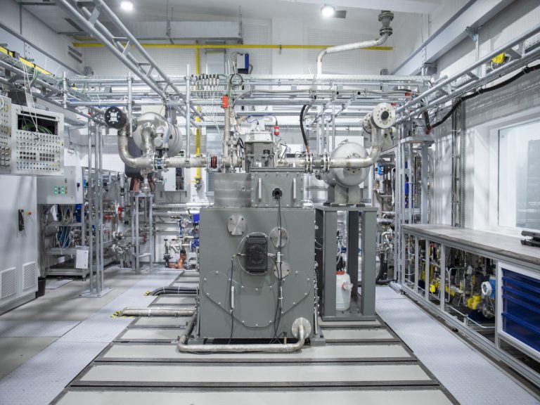 Blick in eine helle Versuchshalle mit einem technisch komplexen Einzylinder-Teststand. Im Zentrum steht eine großformatige Forschungsanlage aus Metall mit zahlreichen Rohrleitungen, Ventilen, Messinstrumenten und Steuerungseinheiten. Die Anlage ist von weiterer Labor- und Prüftechnik umgeben und dient der Untersuchung motorischer Verbrennungsprozesse von Wasserstoff und Ammoniak am WTZ Roßlau.