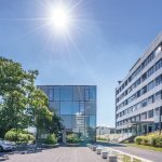 Der gläserne „Energiewürfel“ der Stadtwerke Konstanz steht auf einem modernen Unternehmenscampus. Das kubische Gebäude mit transparenter Glasfassade wird von einem klaren, tiefblauen Himmel und strahlendem Sonnenschein überragt. Rechts befindet sich ein mehrgeschossiges Bürogebäude, links rahmen Bäume und ein Carport den Vorplatz ein. Vor dem Energiewürfel führen Wasserspiele und bepflanzte Sitzbereiche zum Eingangsbereich und unterstreichen den modernen, nachhaltigen Charakter des Standorts.