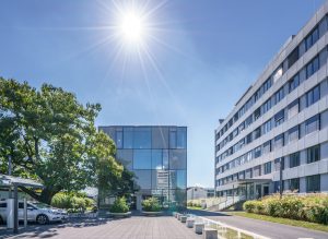 Der gläserne „Energiewürfel“ der Stadtwerke Konstanz steht auf einem modernen Unternehmenscampus. Das kubische Gebäude mit transparenter Glasfassade wird von einem klaren, tiefblauen Himmel und strahlendem Sonnenschein überragt. Rechts befindet sich ein mehrgeschossiges Bürogebäude, links rahmen Bäume und ein Carport den Vorplatz ein. Vor dem Energiewürfel führen Wasserspiele und bepflanzte Sitzbereiche zum Eingangsbereich und unterstreichen den modernen, nachhaltigen Charakter des Standorts.