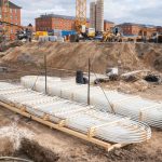 Blick über die Baustelle des Quartiers Lagarde auf dem ehemaligen Kasernengelände in Bamberg. Im Vordergrund liegen gebündelte Kunststoffrohre für die Wärmeversorgung, dahinter sind Bewehrungsarbeiten, Baugruben und Erdarbeiten zu sehen. Mehrere Kräne, Bagger und Baustellenfahrzeuge prägen das Bild. Im Hintergrund stehen sanierte historische Backsteingebäude des entstehenden Campus, der im Rahmen der Wärmewende mit einer klimafreundlichen Energieinfrastruktur ausgestattet wird.