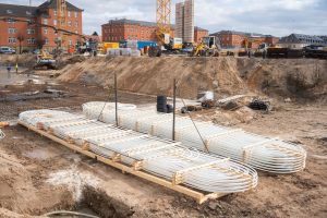 Blick über die Baustelle des Quartiers Lagarde auf dem ehemaligen Kasernengelände in Bamberg. Im Vordergrund liegen gebündelte Kunststoffrohre für die Wärmeversorgung, dahinter sind Bewehrungsarbeiten, Baugruben und Erdarbeiten zu sehen. Mehrere Kräne, Bagger und Baustellenfahrzeuge prägen das Bild. Im Hintergrund stehen sanierte historische Backsteingebäude des entstehenden Campus, der im Rahmen der Wärmewende mit einer klimafreundlichen Energieinfrastruktur ausgestattet wird.