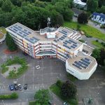 Luftaufnahme des Rathauses von Herzogenrath. Das mehrflügelige Verwaltungsgebäude ist von Bäumen und Grünflächen umgeben. Auf mehreren Flachdächern sind Photovoltaikanlagen installiert, ein Gebäudeteil verfügt über eine extensive Dachbegrünung. Vor dem Haupteingang befinden sich ein großer Vorplatz, Parkplätze und Zufahrtsstraßen.