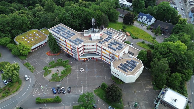 Luftaufnahme des Rathauses von Herzogenrath. Das mehrflügelige Verwaltungsgebäude ist von Bäumen und Grünflächen umgeben. Auf mehreren Flachdächern sind Photovoltaikanlagen installiert, ein Gebäudeteil verfügt über eine extensive Dachbegrünung. Vor dem Haupteingang befinden sich ein großer Vorplatz, Parkplätze und Zufahrtsstraßen.