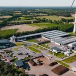 Luftaufnahme eines Energie- und Kompostwerks in Saerbeck. Zu sehen sind mehrere Hallen, eine Biogasanlage mit technischen Anlagen und Rohrleitungen, Lagerflächen sowie ein Biomassebereich. Im Hintergrund erstrecken sich Felder und Waldflächen, daneben steht eine Windenergieanlage.