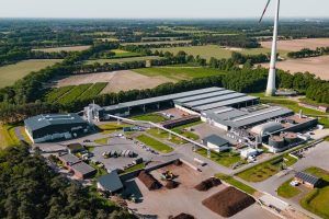Luftaufnahme eines Energie- und Kompostwerks in Saerbeck. Zu sehen sind mehrere Hallen, eine Biogasanlage mit technischen Anlagen und Rohrleitungen, Lagerflächen sowie ein Biomassebereich. Im Hintergrund erstrecken sich Felder und Waldflächen, daneben steht eine Windenergieanlage.