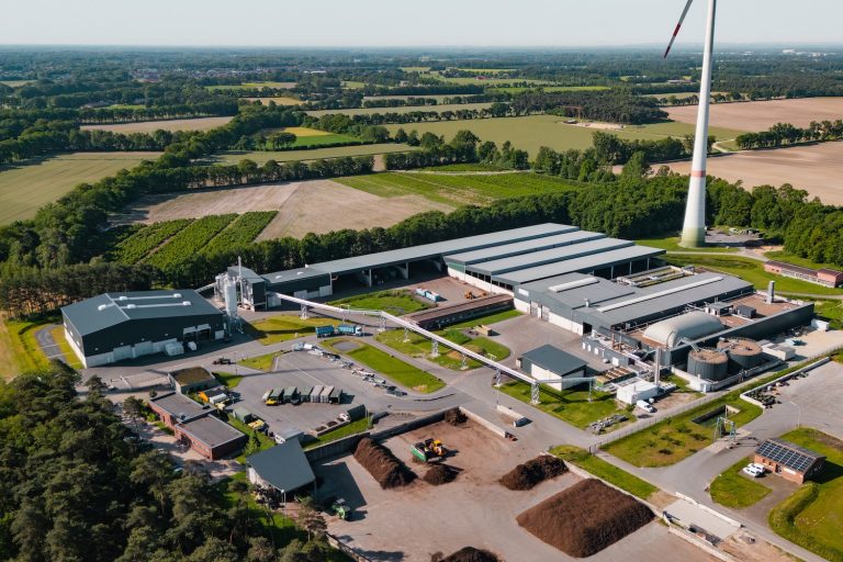 Luftaufnahme eines Energie- und Kompostwerks in Saerbeck. Zu sehen sind mehrere Hallen, eine Biogasanlage mit technischen Anlagen und Rohrleitungen, Lagerflächen sowie ein Biomassebereich. Im Hintergrund erstrecken sich Felder und Waldflächen, daneben steht eine Windenergieanlage.