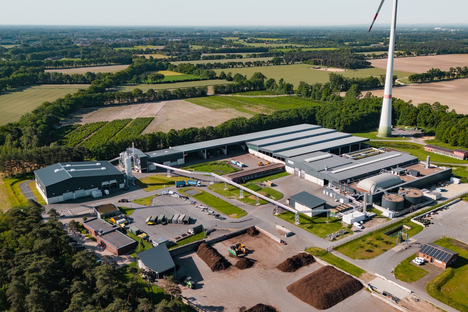 Luftaufnahme eines Energie- und Kompostwerks in Saerbeck. Zu sehen sind mehrere Hallen, eine Biogasanlage mit technischen Anlagen und Rohrleitungen, Lagerflächen sowie ein Biomassebereich. Im Hintergrund erstrecken sich Felder und Waldflächen, daneben steht eine Windenergieanlage.