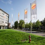 Modernes Bürogebäude des Energieversorgers Entega mit mehreren Fahnenmasten, auf denen Entega-Flaggen wehen. Vor dem Gebäude befinden sich eine gepflegte Rasenfläche, ein Parkplatz mit Fahrrädern und Autos sowie Bäume. Der Himmel ist klar und sonnig.