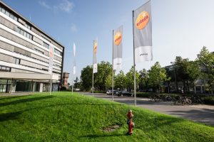 Modernes Bürogebäude des Energieversorgers Entega mit mehreren Fahnenmasten, auf denen Entega-Flaggen wehen. Vor dem Gebäude befinden sich eine gepflegte Rasenfläche, ein Parkplatz mit Fahrrädern und Autos sowie Bäume. Der Himmel ist klar und sonnig.