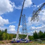 Ein großer Mobilkran richtet auf einer Freifläche am Waldrand einen Gittermast einer Hochspannungsfreileitung auf. Mehrere Monteure begleiten die Montage, während weitere Mastsegmente am Boden bereitliegen. Über der Baustelle erstreckt sich ein blauer Himmel mit großen weißen Wolken.