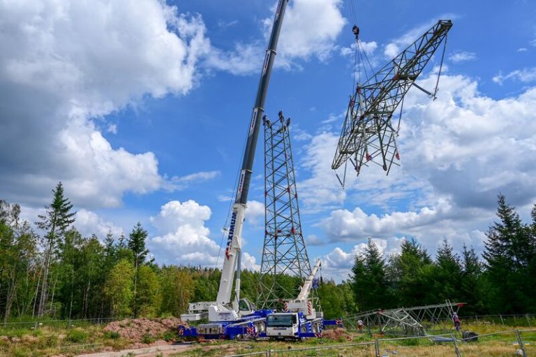Ein großer Mobilkran richtet auf einer Freifläche am Waldrand einen Gittermast einer Hochspannungsfreileitung auf. Mehrere Monteure begleiten die Montage, während weitere Mastsegmente am Boden bereitliegen. Über der Baustelle erstreckt sich ein blauer Himmel mit großen weißen Wolken.